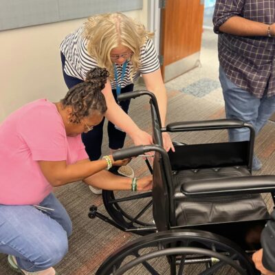 Two participants work together to assemble a wheelchair during the Wheelchairs In Motion team building event. They focus on adjusting and securing parts of the wheelchair frame as part of the charitable activity. thumbnail