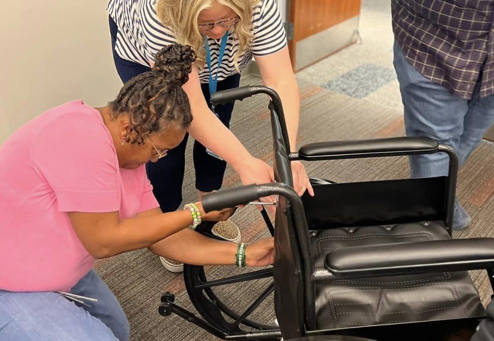Two participants work together to assemble a wheelchair during the Wheelchairs In Motion team building event. They focus on adjusting and securing parts of the wheelchair frame as part of the charitable activity.