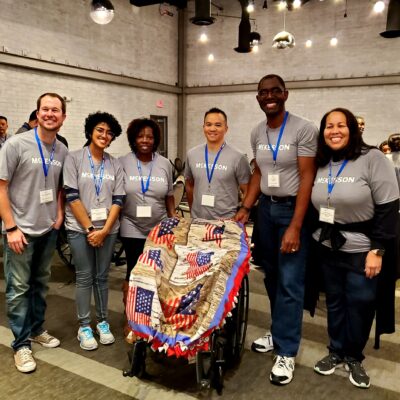 A group of participants stands together smiling, next to a wheelchair decorated with a patriotic blanket during the Wheelchairs In Motion team building event. The team wears matching event shirts and badges, celebrating their successful charitable effort. thumbnail