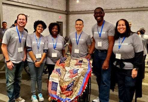 A group of participants stands together smiling, next to a wheelchair decorated with a patriotic blanket during the Wheelchairs In Motion team building event. The team wears matching event shirts and badges, celebrating their successful charitable effort.