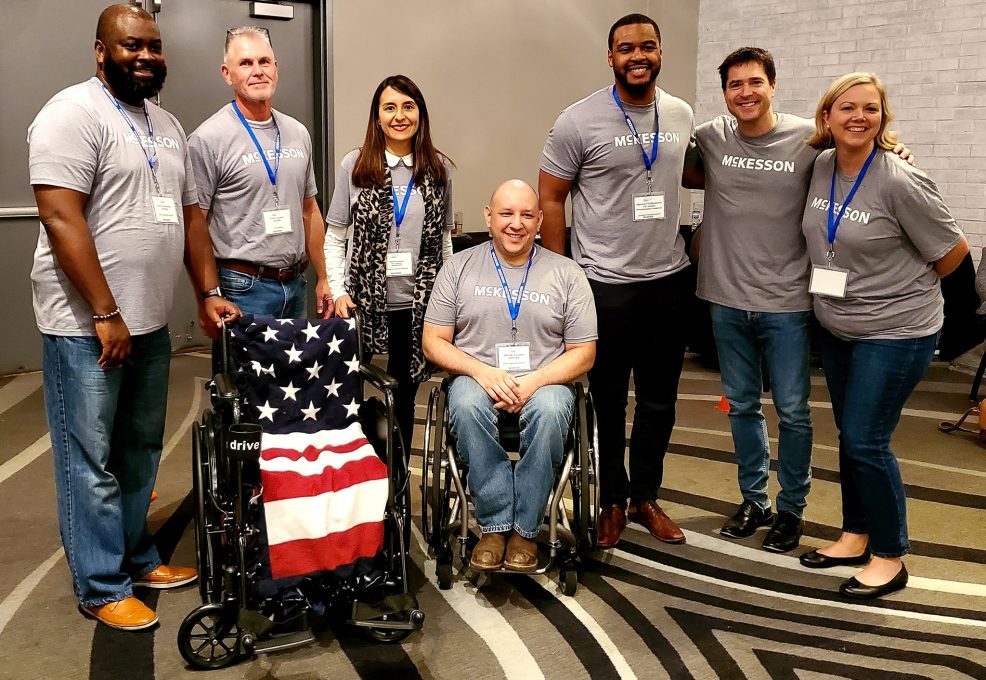 A group of participants poses together, smiling, at the Wheelchairs In Motion team building event. One participant is seated in a wheelchair next to another decorated with an American flag blanket, showcasing their involvement in the charitable activity.