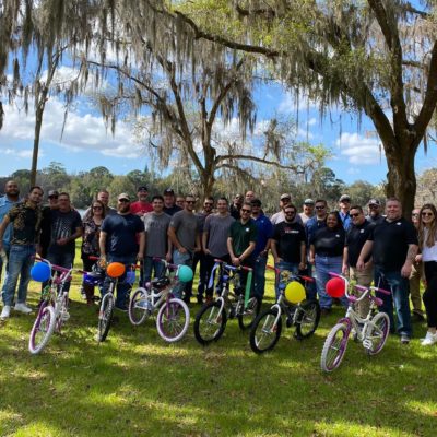 Large group of participants posing outdoors with completed bikes from a Charity Bike Build event. Each assembled bike is decorated with colorful balloons. Participants are smiling in a celebratory mood after a successful team building activity. thumbnail