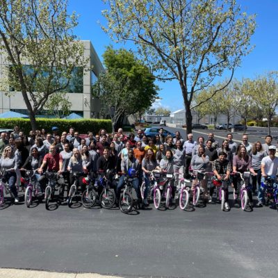 Large group of participants posing outdoors with assembled bikes during a Charity Bike Build event. The group stands proudly behind the rows of bikes, enjoying the sunny day and the sense of accomplishment after working together for a charitable cause. thumbnail