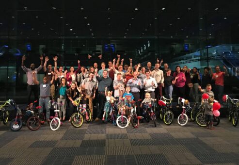 Group of participants smiling and cheering indoors during a Charity Bike Build event. In the front, children excitedly stand with their newly built bikes, surrounded by colorful balloons. The event celebrates teamwork and community engagement as the group successfully completes the charity project.