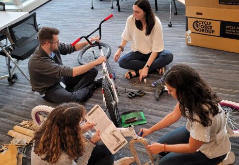 A group of four people sitting on the floor assembling a bicycle during a Charity Bike Build event. They are carefully reading instructions and working collaboratively, with bike parts and tools spread around them.