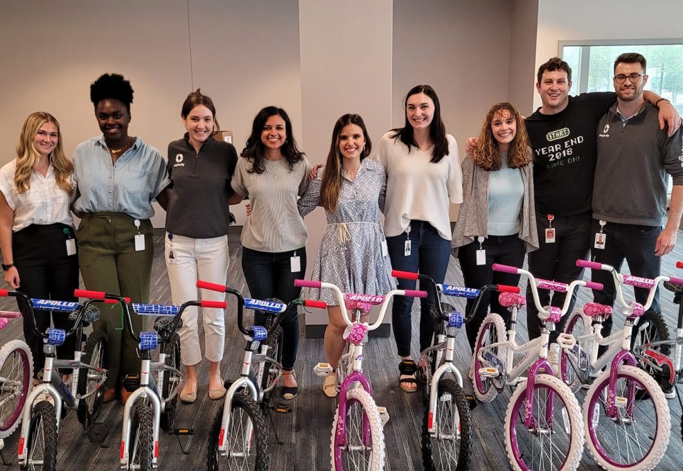A group of smiling participants standing behind a row of assembled children's bicycles during a Charity Bike Build event. The bikes are pink and blue with training wheels, and the group appears happy with their completed team project.