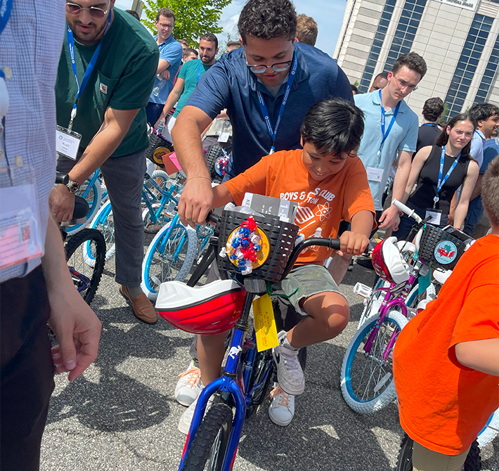 A group of volunteers assisting children in riding their newly assembled bikes during a Charity Bike Build event. A boy in an orange shirt excitedly rides a decorated blue bike, guided by a volunteer.