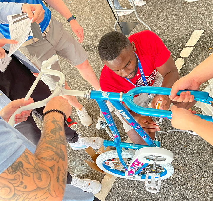 A group of participants working together to assemble a children's bicycle during a Charity Bike Build event. One person is tightening a bolt while others hold the bike steady and refer to instructions, demonstrating teamwork and collaboration.