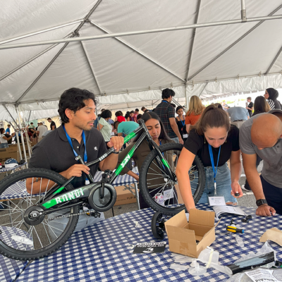 A group of participants working together to assemble a green bicycle during a Charity Bike Build event. They are under a large tent, focused on attaching parts with tools. Other groups in the background are also working on assembling bikes. thumbnail