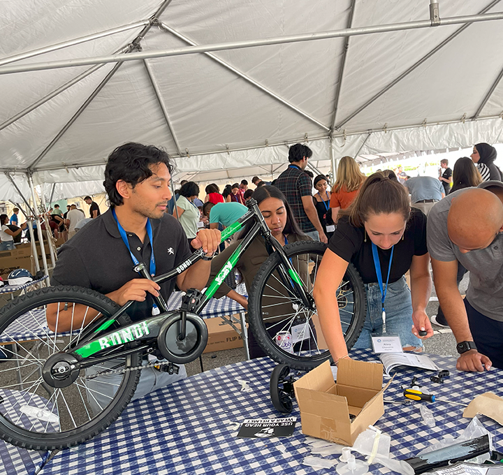 A group of participants working together to assemble a green bicycle during a Charity Bike Build event. They are under a large tent, focused on attaching parts with tools. Other groups in the background are also working on assembling bikes.