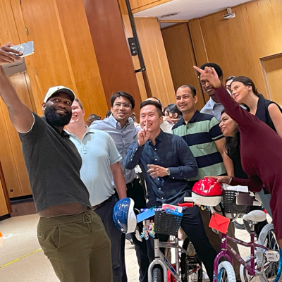 Group of enthusiastic participants taking a selfie during a Charity Bike Build event. They are smiling and posing with peace signs while standing next to the bicycles they assembled, which are adorned with helmets and decorations, highlighting the fun and collaborative atmosphere of the event. thumbnail