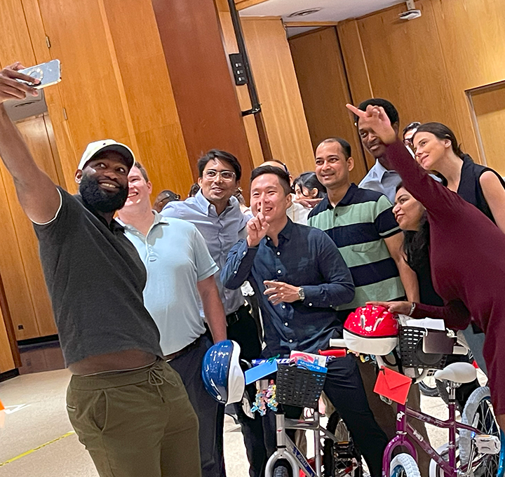 Group of enthusiastic participants taking a selfie during a Charity Bike Build event. They are smiling and posing with peace signs while standing next to the bicycles they assembled, which are adorned with helmets and decorations, highlighting the fun and collaborative atmosphere of the event.