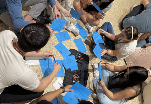 A group of people sitting in a circle on the floor during a team building activity, each holding blue cards with instructions. They are engaging in a collaborative task, exchanging ideas and writing down notes.