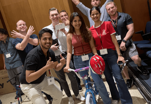 A group of smiling participants posing proudly with a completed bicycle during a Charity Bike Build event. Some team members are giving thumbs up and raising their hands in excitement, celebrating their achievement after assembling the bike.