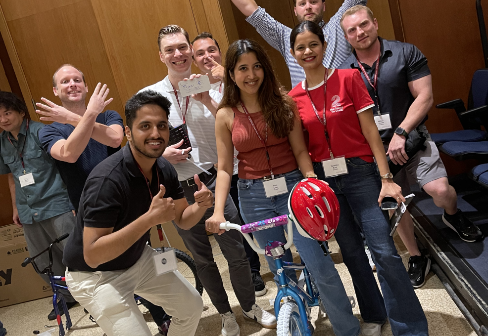 A group of smiling participants posing proudly with a completed bicycle during a Charity Bike Build event. Some team members are giving thumbs up and raising their hands in excitement, celebrating their achievement after assembling the bike.