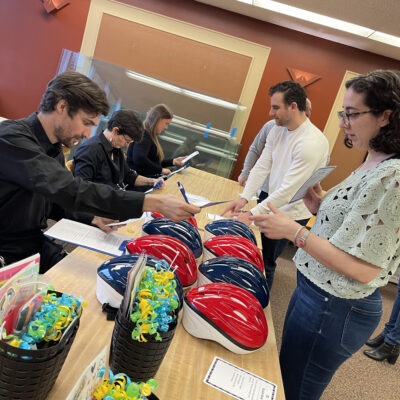 Participants in a team building event stand at a table with red and blue bicycle helmets, discussing instructions and checking documents. Gift baskets with colorful decorations are also displayed, highlighting the preparation phase of a Charity Bike Build event. thumbnail