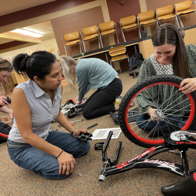 Four team members work together on the floor to assemble bikes during the Charity Bike Build event. They are focused on attaching wheels and following instructions, demonstrating collaboration and problem-solving as part of this hands-on, team building activity. thumbnail