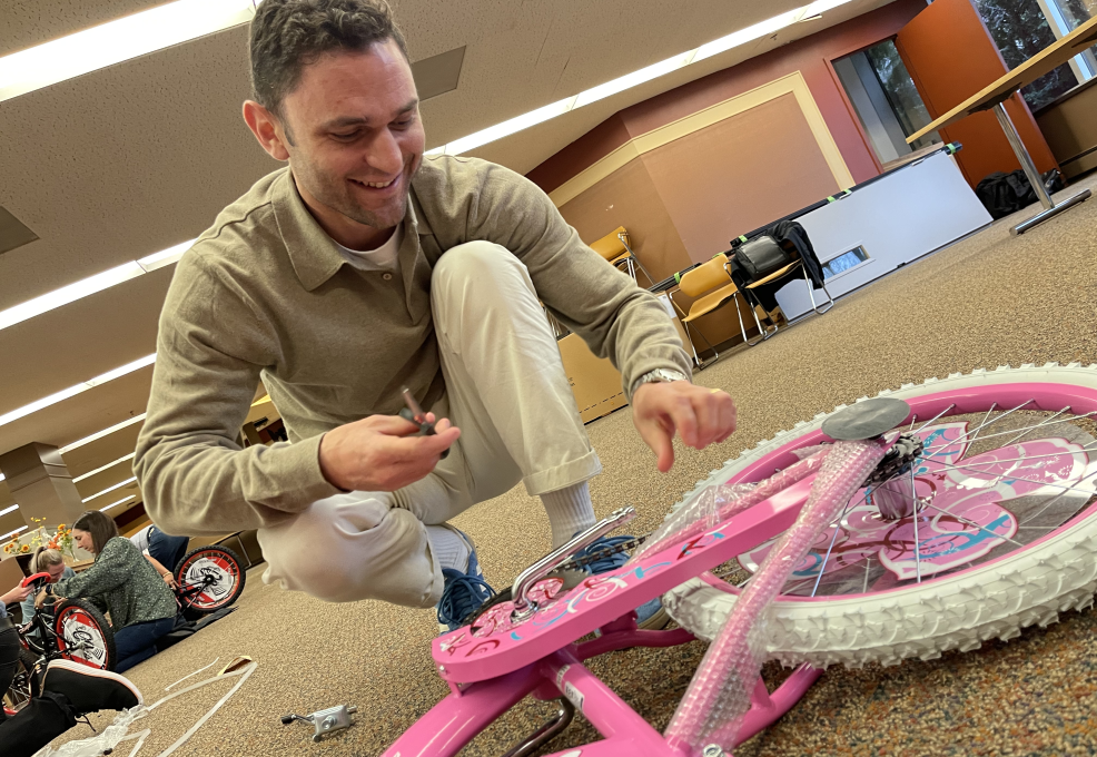 A man crouches on the floor with a smile, carefully assembling a pink bicycle during a Charity Bike Build team building event. He uses tools to attach the final pieces, showcasing hands-on involvement in creating bikes for children.
