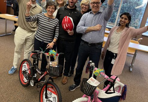A group of seven participants cheer enthusiastically while standing behind two completed bikes during a Charity Bike Build event.