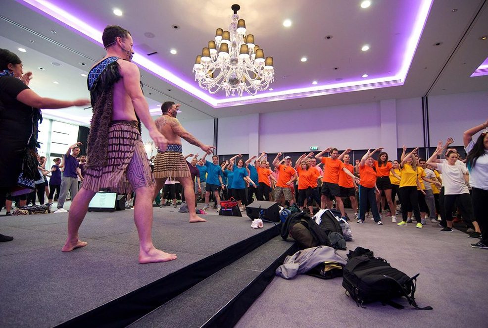 Maori performers in traditional attire leading a Haka team building session for a large group of participants.