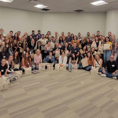A large group of participants pose for a group photo at the conclusion of a team building cooking activity. The group, consisting of over 50 people, is smiling and seated on the floor of a spacious room. Many participants are holding decorated bags, filled with food donations they prepared during the event. The atmosphere appears to be joyful and celebratory, marking the success of their collaborative efforts to give back to the community. thumbnail