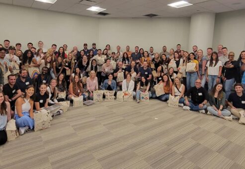 A large group of participants pose for a group photo at the conclusion of a team building cooking activity. The group, consisting of over 50 people, is smiling and seated on the floor of a spacious room. Many participants are holding decorated bags, filled with food donations they prepared during the event. The atmosphere appears to be joyful and celebratory, marking the success of their collaborative efforts to give back to the community.