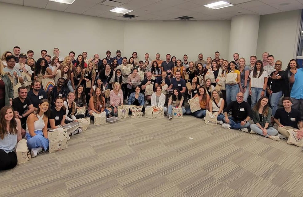 A large group of participants pose for a group photo at the conclusion of a team building cooking activity. The group, consisting of over 50 people, is smiling and seated on the floor of a spacious room. Many participants are holding decorated bags, filled with food donations they prepared during the event. The atmosphere appears to be joyful and celebratory, marking the success of their collaborative efforts to give back to the community.