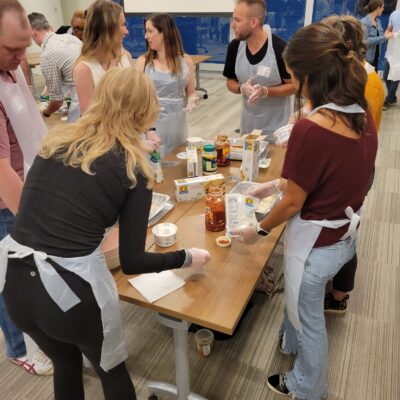 A group of participants wearing aprons and gloves are engaged in a team building cooking activity. They are gathered around a table, preparing ingredients like pasta sauce and cheese to assemble lasagna dishes. The group is conversing and collaborating, with a variety of food items and kitchen tools spread out on the table. The setting appears lively and focused as they work together to prepare meals for donation. thumbnail