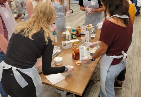 A group of participants wearing aprons and gloves are engaged in a team building cooking activity. They are gathered around a table, preparing ingredients like pasta sauce and cheese to assemble lasagna dishes. The group is conversing and collaborating, with a variety of food items and kitchen tools spread out on the table. The setting appears lively and focused as they work together to prepare meals for donation.