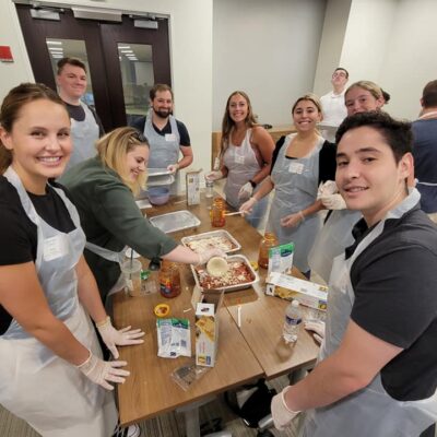 A group of nine individuals is gathered around a table, all smiling and wearing aprons and gloves as they participate in a team building cooking activity. One person is scooping cheese onto a lasagna dish, while various other ingredients like sauce, pasta, and cheese are arranged on the table. The group looks happy and engaged in their task as they prepare meals together. thumbnail