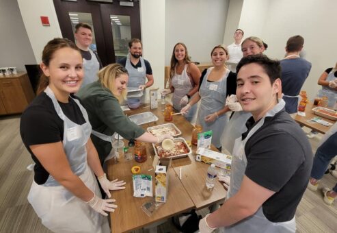 A group of nine individuals is gathered around a table, all smiling and wearing aprons and gloves as they participate in a team building cooking activity. One person is scooping cheese onto a lasagna dish, while various other ingredients like sauce, pasta, and cheese are arranged on the table. The group looks happy and engaged in their task as they prepare meals together.