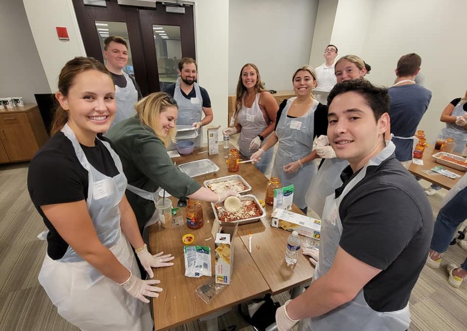 A group of nine individuals is gathered around a table, all smiling and wearing aprons and gloves as they participate in a team building cooking activity. One person is scooping cheese onto a lasagna dish, while various other ingredients like sauce, pasta, and cheese are arranged on the table. The group looks happy and engaged in their task as they prepare meals together.