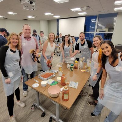 A group of eight people stands around a table, smiling for the camera while participating in a team building cooking activity. Each person is wearing an apron and gloves, and various ingredients such as pasta, sauce, and cheese are spread out on the table. Some of the participants are holding jars of sauce, ready to assemble a dish. The group is engaged in preparing meals as part of a community service project. thumbnail