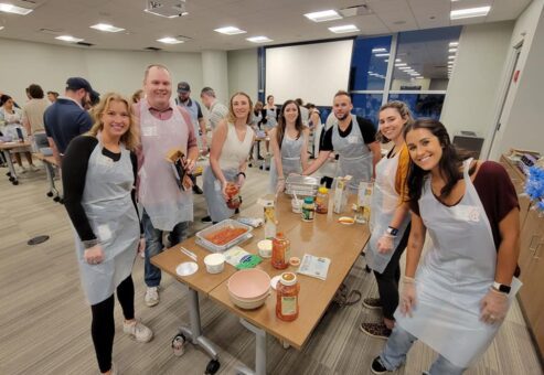 A group of eight people stands around a table, smiling for the camera while participating in a team building cooking activity. Each person is wearing an apron and gloves, and various ingredients such as pasta, sauce, and cheese are spread out on the table. Some of the participants are holding jars of sauce, ready to assemble a dish. The group is engaged in preparing meals as part of a community service project.
