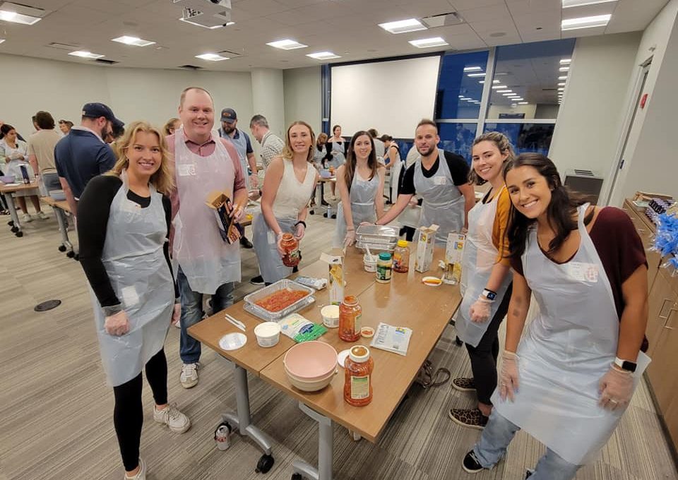 A group of eight people stands around a table, smiling for the camera while participating in a team building cooking activity. Each person is wearing an apron and gloves, and various ingredients such as pasta, sauce, and cheese are spread out on the table. Some of the participants are holding jars of sauce, ready to assemble a dish. The group is engaged in preparing meals as part of a community service project.
