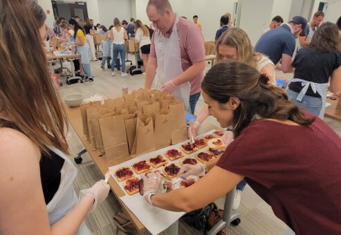 A group of people participating in a team building activity are preparing sandwiches on a large table. The participants, wearing aprons and gloves, are spreading peanut butter and jelly on slices of bread. In the background, more individuals are working at various tables, all focused on assembling sandwiches. On the table, a row of brown paper bags is lined up, ready to be filled with the prepared sandwiches. The room is filled with activity as people work together to contribute to a food donation project.