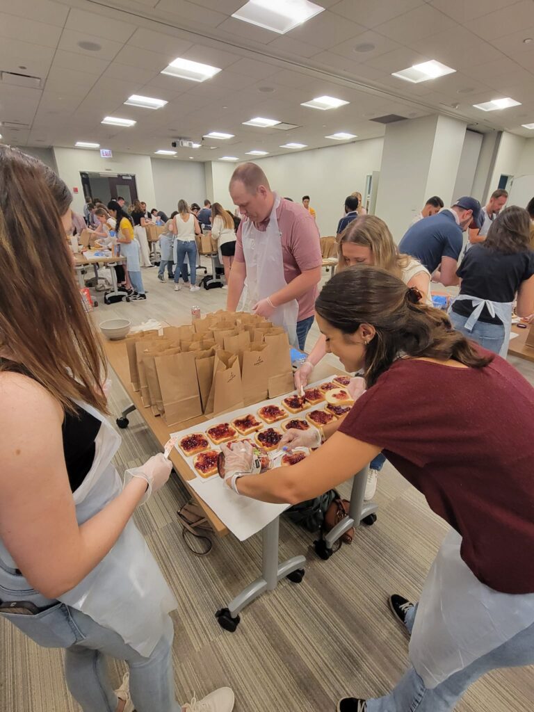 A group of people participating in a team building activity are preparing sandwiches on a large table. The participants, wearing aprons and gloves, are spreading peanut butter and jelly on slices of bread. In the background, more individuals are working at various tables, all focused on assembling sandwiches. On the table, a row of brown paper bags is lined up, ready to be filled with the prepared sandwiches. The room is filled with activity as people work together to contribute to a food donation project.