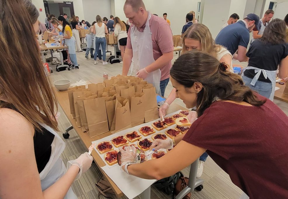 A group of people participating in a team building activity are preparing sandwiches on a large table. The participants, wearing aprons and gloves, are spreading peanut butter and jelly on slices of bread. In the background, more individuals are working at various tables, all focused on assembling sandwiches. On the table, a row of brown paper bags is lined up, ready to be filled with the prepared sandwiches. The room is filled with activity as people work together to contribute to a food donation project.