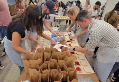 Participants wearing aprons and gloves are working together in a large room to prepare sandwiches for donation. They are spreading peanut butter and jelly on slices of bread laid out on a long table. In the foreground, brown paper bags are lined up, ready to be filled with sandwiches. The room is bustling with activity as several groups of people are engaged in similar tasks at other tables, contributing to a CSR team building program.
