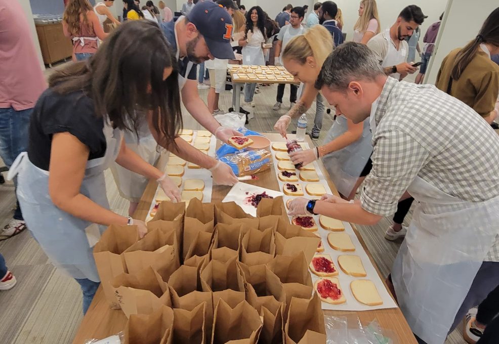 Participants wearing aprons and gloves are working together in a large room to prepare sandwiches for donation. They are spreading peanut butter and jelly on slices of bread laid out on a long table. In the foreground, brown paper bags are lined up, ready to be filled with sandwiches. The room is bustling with activity as several groups of people are engaged in similar tasks at other tables, contributing to a CSR team building program.