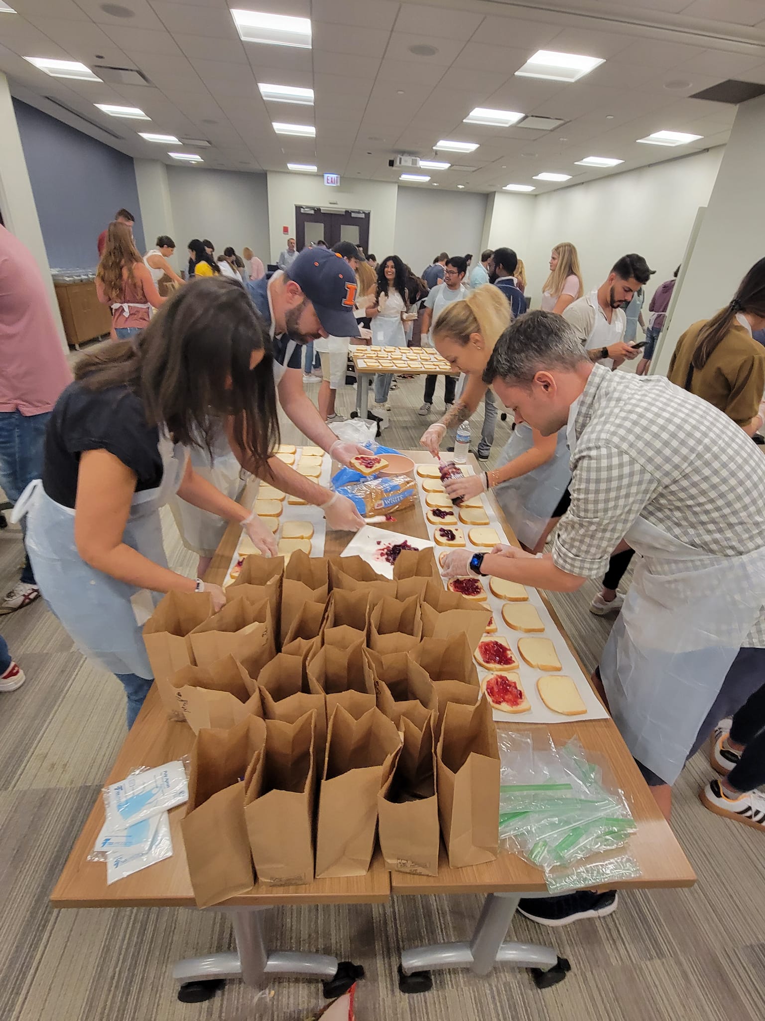 Participants wearing aprons and gloves are working together in a large room to prepare sandwiches for donation. They are spreading peanut butter and jelly on slices of bread laid out on a long table. In the foreground, brown paper bags are lined up, ready to be filled with sandwiches. The room is bustling with activity as several groups of people are engaged in similar tasks at other tables, contributing to a CSR team building program.