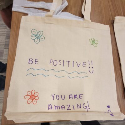 A decorated canvas tote bag lays flat on a wooden table. The bag features hand-drawn flowers in various colors and motivational phrases written in colorful markers. The text on the bag reads 'BE POSITIVE!!' with a smiley face, followed by 'YOU ARE AMAZING!' below. The design is simple and cheerful, meant to inspire and encourage. thumbnail