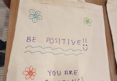 A decorated canvas tote bag lays flat on a wooden table. The bag features hand-drawn flowers in various colors and motivational phrases written in colorful markers. The text on the bag reads 'BE POSITIVE!!' with a smiley face, followed by 'YOU ARE AMAZING!' below. The design is simple and cheerful, meant to inspire and encourage.