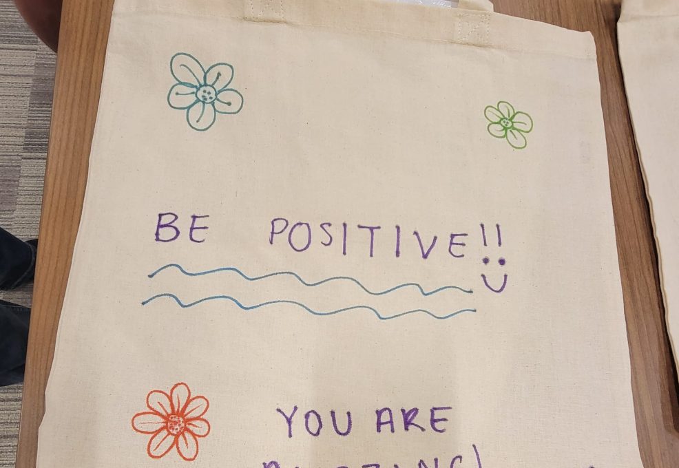 A decorated canvas tote bag lays flat on a wooden table. The bag features hand-drawn flowers in various colors and motivational phrases written in colorful markers. The text on the bag reads 'BE POSITIVE!!' with a smiley face, followed by 'YOU ARE AMAZING!' below. The design is simple and cheerful, meant to inspire and encourage.