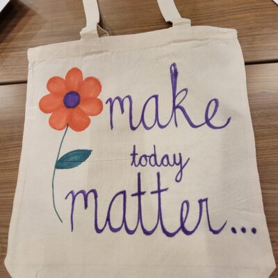 A beige fabric tote bag displayed on a wooden table, decorated with a hand-painted orange flower with a purple center and green stem. Next to the flower, the words 'make today matter...' are written in purple cursive, encouraging a positive message. The bag is part of a CSR team building event.