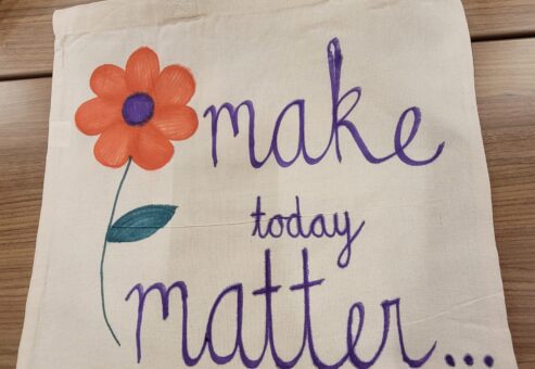 A beige fabric tote bag displayed on a wooden table, decorated with a hand-painted orange flower with a purple center and green stem. Next to the flower, the words 'make today matter...' are written in purple cursive, encouraging a positive message. The bag is part of a CSR team building event.