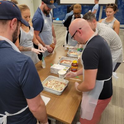 A group of participants wearing aprons and gloves are gathered around a table, preparing trays of pasta for donation. They are adding ingredients like sauce and cheese to large aluminum trays. The participants are engaged and working together in a well-lit room, with various ingredients and supplies on the table. This is part of a CSR team building event aimed at preparing meals for those in need. thumbnail