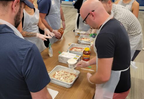 A group of participants wearing aprons and gloves are gathered around a table, preparing trays of pasta for donation. They are adding ingredients like sauce and cheese to large aluminum trays. The participants are engaged and working together in a well-lit room, with various ingredients and supplies on the table. This is part of a CSR team building event aimed at preparing meals for those in need.