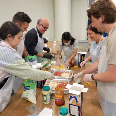 A group of six participants, wearing aprons and gloves, are gathered around a table during a team building cooking activity. They are preparing a lasagna dish using ingredients such as pasta sauce, cheese, and lasagna sheets. The group appears cheerful and collaborative as they layer ingredients into trays. Various jars, boxes, and containers are scattered on the table as they work together to complete the task. thumbnail