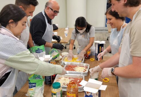 A group of six participants, wearing aprons and gloves, are gathered around a table during a team building cooking activity. They are preparing a lasagna dish using ingredients such as pasta sauce, cheese, and lasagna sheets. The group appears cheerful and collaborative as they layer ingredients into trays. Various jars, boxes, and containers are scattered on the table as they work together to complete the task.